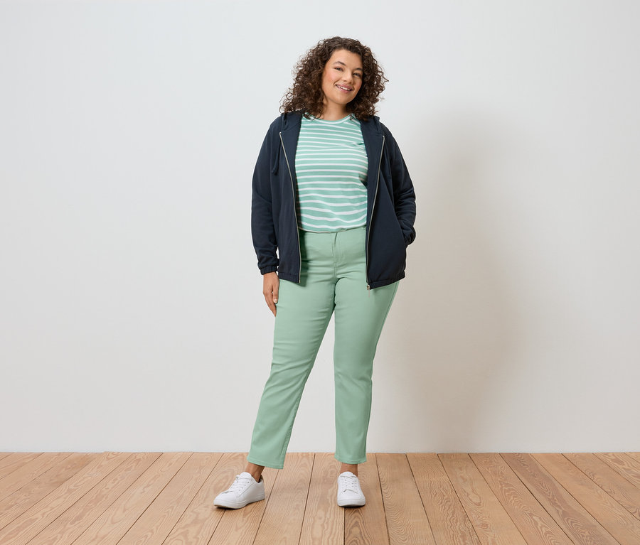 Kobieta z kręconymi włosami stoi na tle ściany, ubrana w granatową kurtkę, koszulkę w paski, zielone spodnie i białe buty.