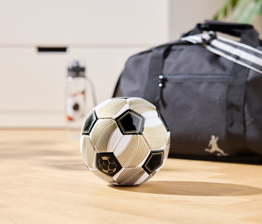 Złota piłka nożna leży na drewnianej podłodze, w tle torba sportowa i butelka na wodę.