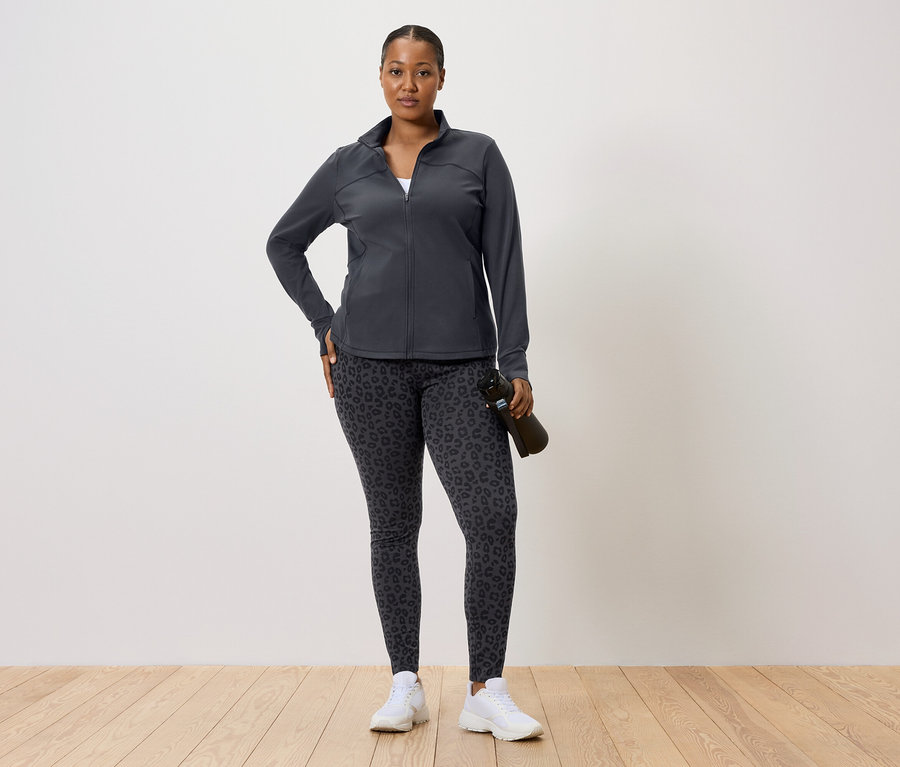 Kobieta w ciemnej kurtce sportowej i legginsach w panterkę, trzyma butelkę z wodą.