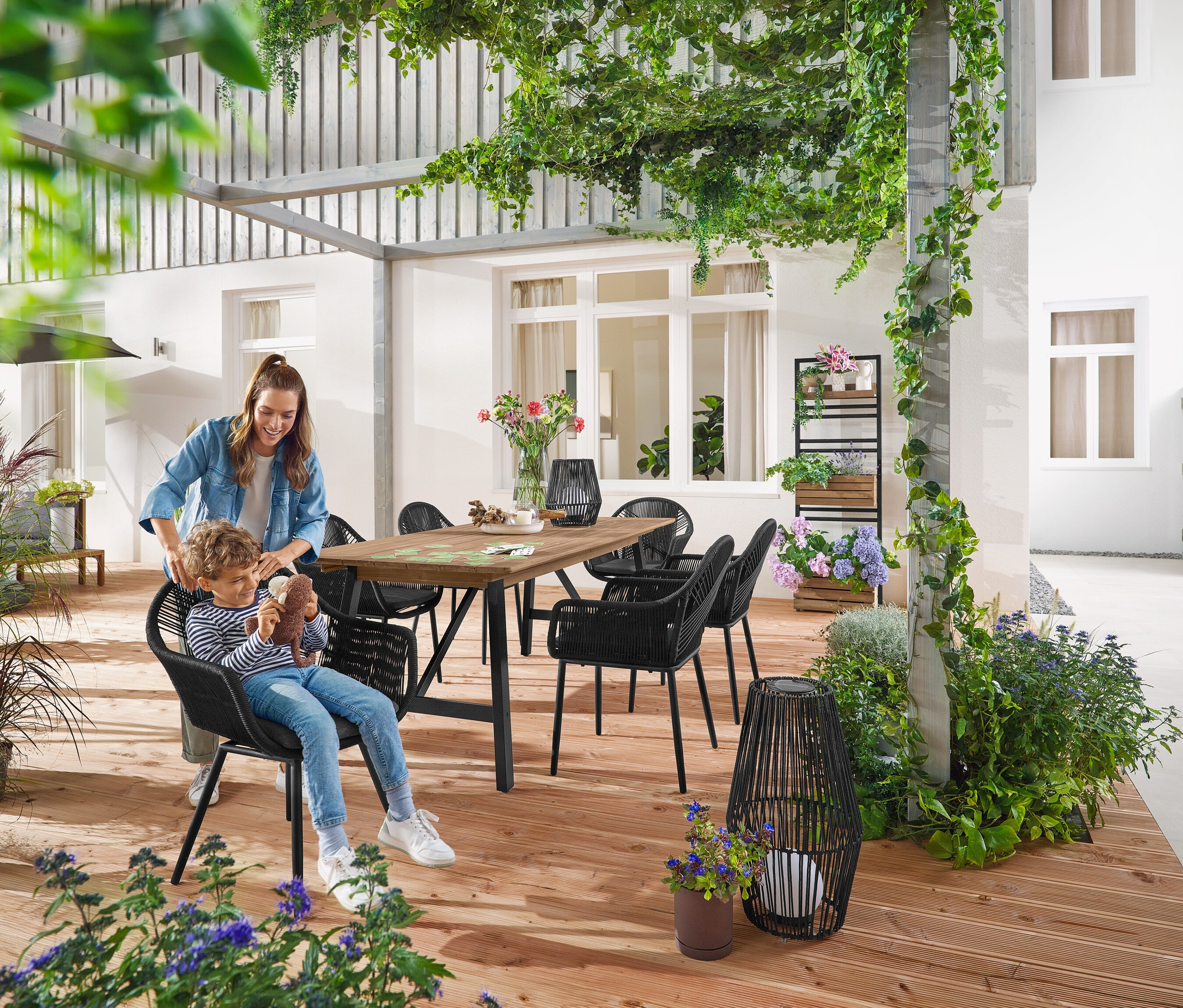 Matka i dziecko siedzą przy Rozkładanym stole ogrodowym o rustykalnej konstrukcji, ok. 200‒250 cm. Mała i duża Latarnia solarna ze sznurkiem tekstylnym stoi na ziemi. W tle Pflanzwand mit flexibel einhängbaren Regalen »Elin«.