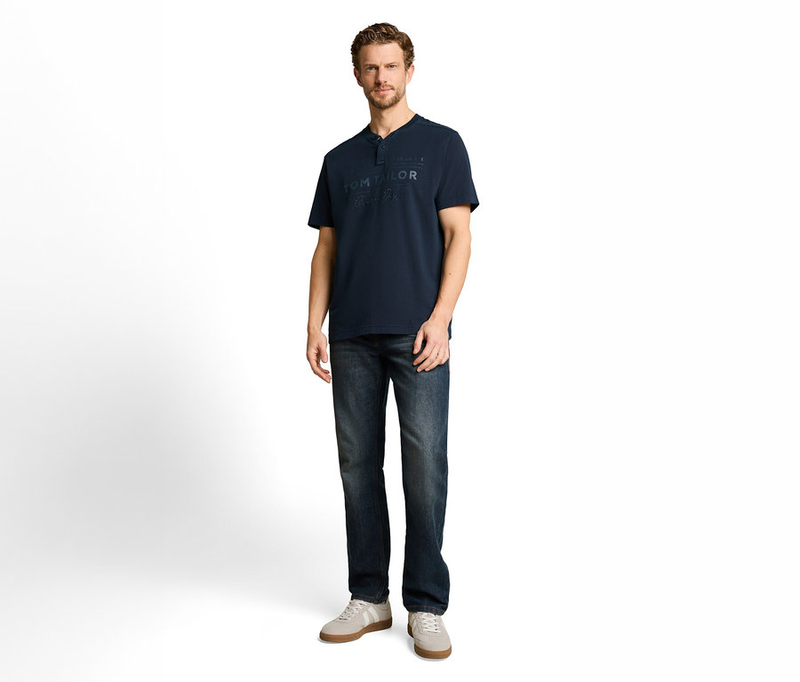 Mężczyzna w granatowej koszulce Henley z logo „TOM TAILOR”, ciemnych dżinsach i jasnoszarych sneakersach.