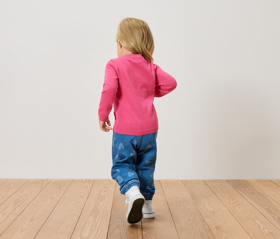 Małe blond dziecko widziane z tyłu, ubrane w różowy sweter, niebieskie dżinsy w serca i białe buty, idzie po drewnianej podłodze.