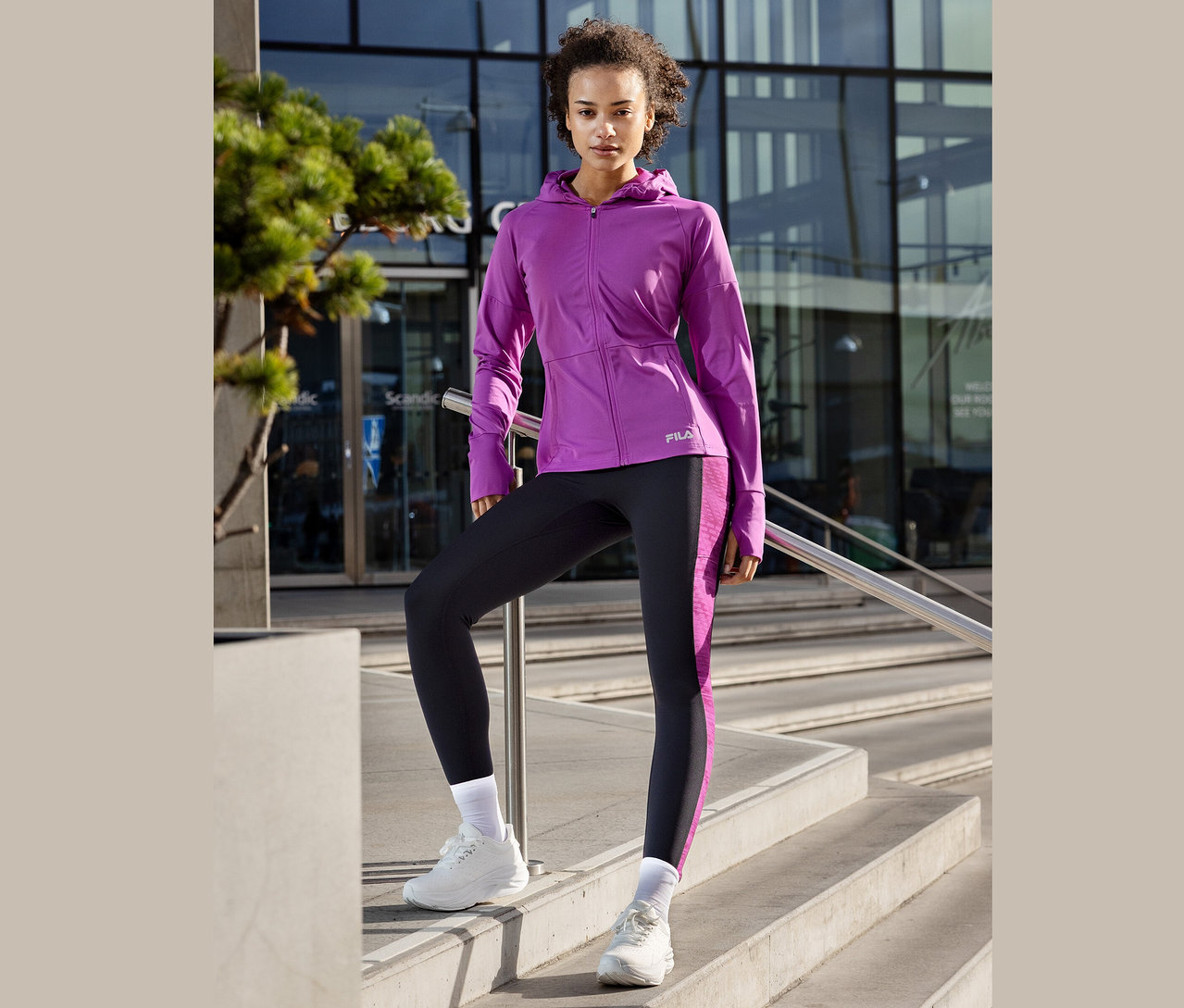 Kobieta pozuje na schodach, ubrana w fioletową kurtkę Fila, legginsy z fioletowymi paskami i białe tenisówki.