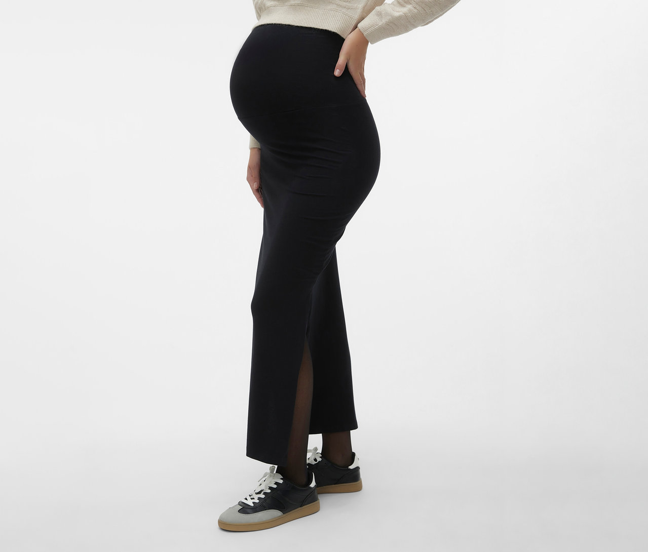 Kobieta w ciąży ma na sobie spódnicę maxi MAMALICIOUS „MLEMMA TUBE” z rozcięciem z boku, ciemne rajstopy i tenisówki na neutralnym tle.
