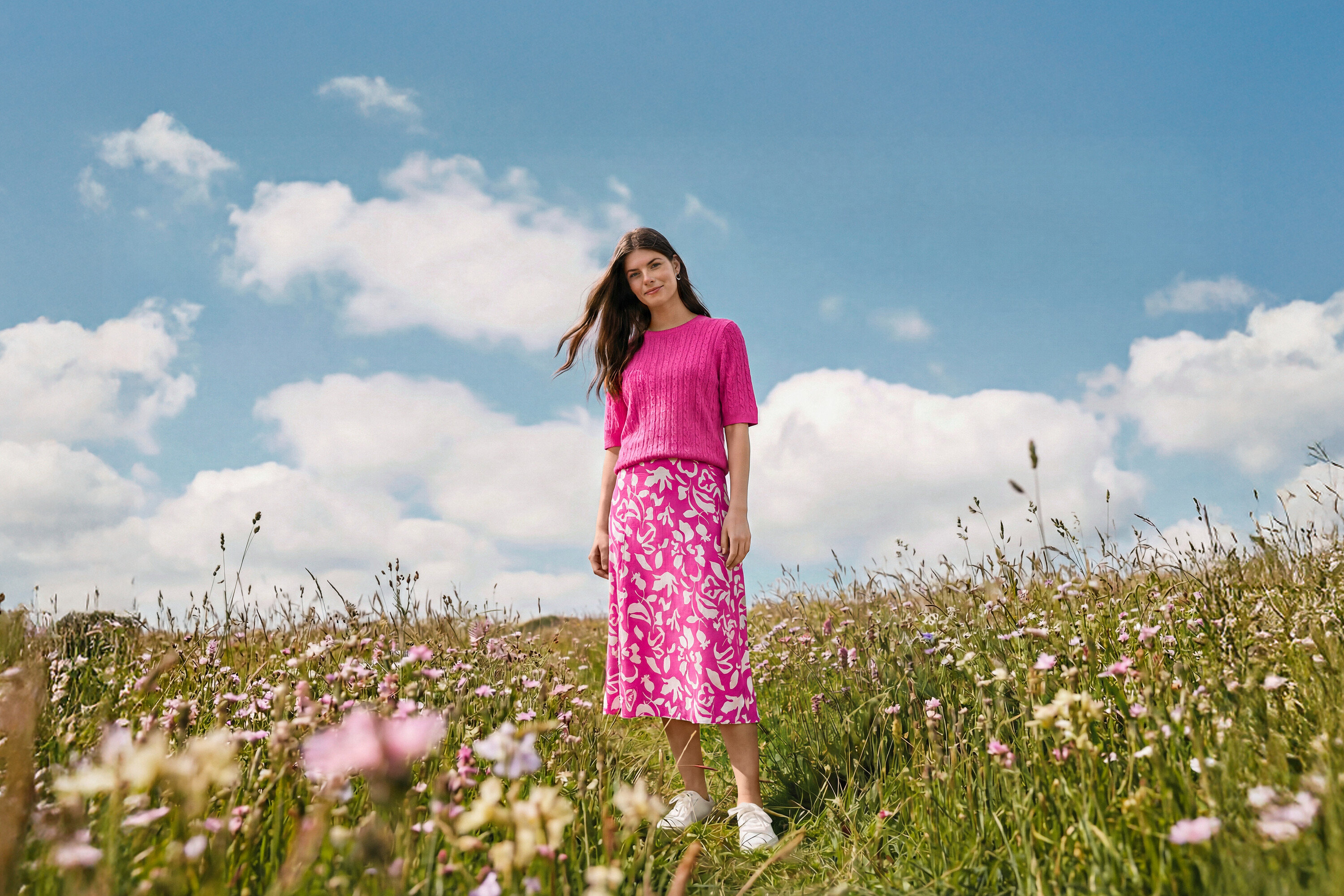 Kobieta stoi na łące w wzorzystej spódnicy midi, kolczykach kreolach Wave i skórzanych sneakersach.