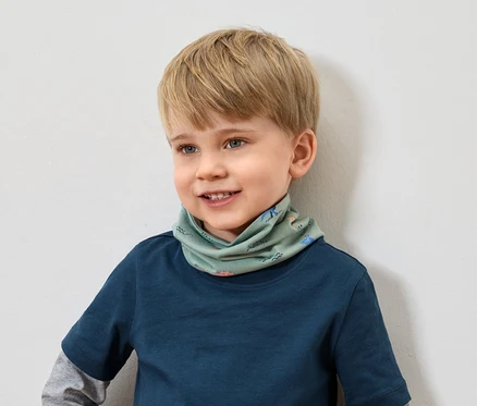 Uśmiechnięty chłopiec z blond włosami w niebieskiej koszulce nosi zielony szalik we wzory zwierzęce.