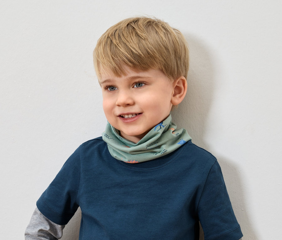 Uśmiechnięty chłopiec z blond włosami w niebieskiej koszulce nosi zielony szalik we wzory zwierzęce.
