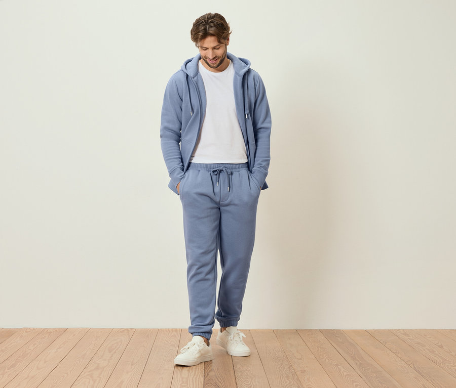 Mężczyzna stoi z rękami w kieszeniach w jasnoniebieskim dresie i białych tenisówkach.