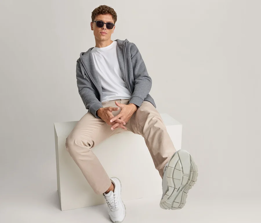 Mężczyzna siedzi na białym bloku, ma na sobie okulary przeciwsłoneczne, biały T-shirt, rozpiętą szarą bluzę z kapturem, jasnobeżowe spodnie i białe sneakersy.