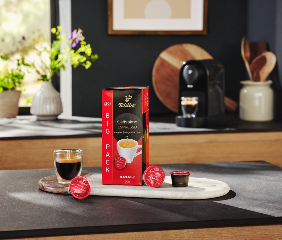 Na stole stoi opakowanie kapsułek Cafissimo Espresso, szklanka espresso i ekspres do kawy.
