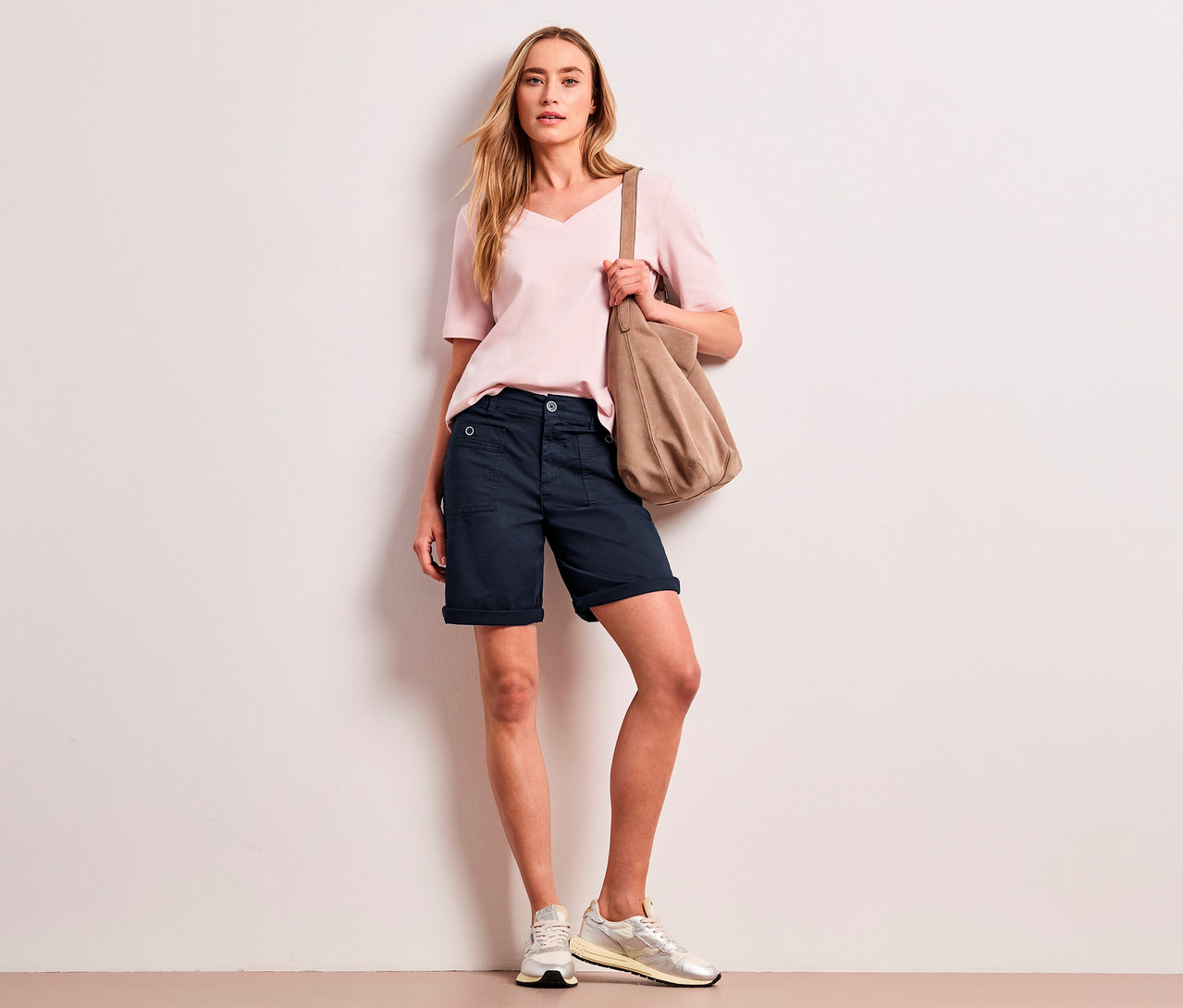 Kobieta z długimi blond włosami ma na sobie jasnoróżowy top, granatowe bermudy Street One „Yulius” i jasne sneakersy, trzymając beżową torbę na ramię.