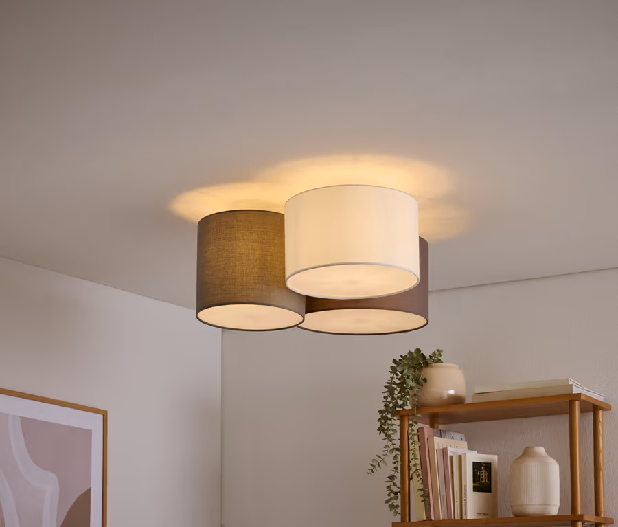 Trzy lampy sufitowe z cylindrycznymi abażurami w kolorach białym, ciemnoszarym i brązowym są zgrupowane na białym suficie. W prawym dolnym rogu widać drewnianą półkę z książkami i rośliną, a w lewym dolnym rogu oprawiony abstrakcyjny obraz.
