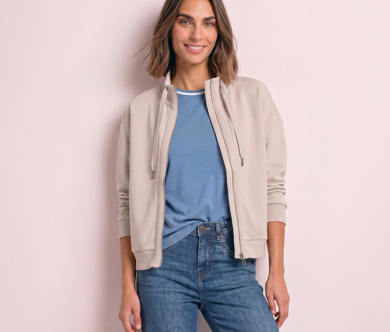 Uśmiechnięta kobieta w beżowej, rozpiętej bluzie dresowej Street One, niebieskim T-shircie i jeansach.