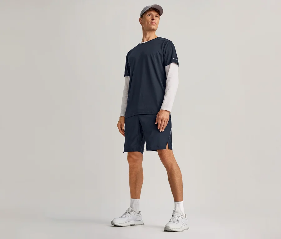 Mężczyzna w granatowych szortach sportowych, granatowej koszulce na białej koszulce z długim rękawem, białych skarpetkach, białych sneakersach i szarej czapce.