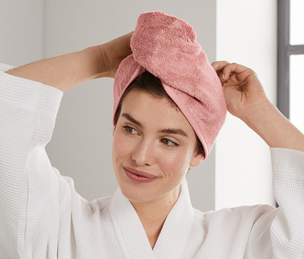 Kobieta poprawia różowy ręcznik-turban na głowie. Ubrana w biały szlafrok stoi w łazience.