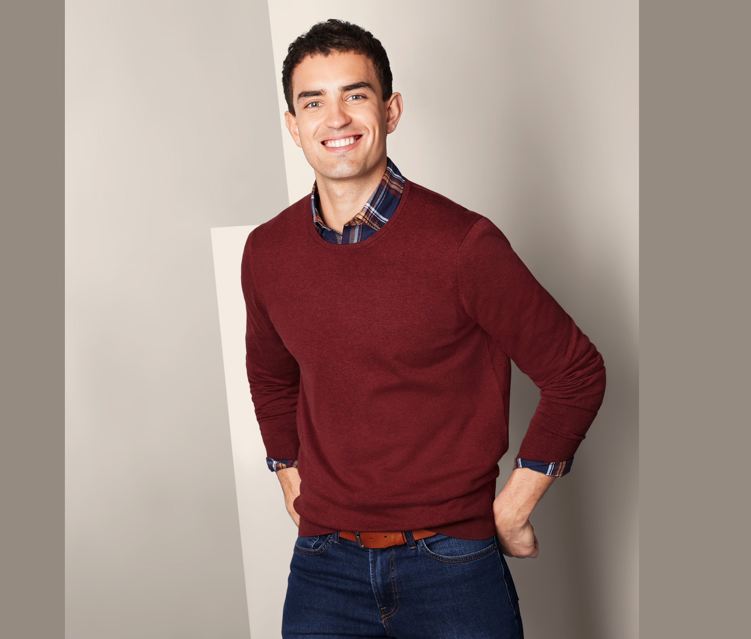 Uśmiechnięty mężczyzna pozuje w bordowym swetrze z okrągłym dekoltem, dżinsach i brązowym pasku.