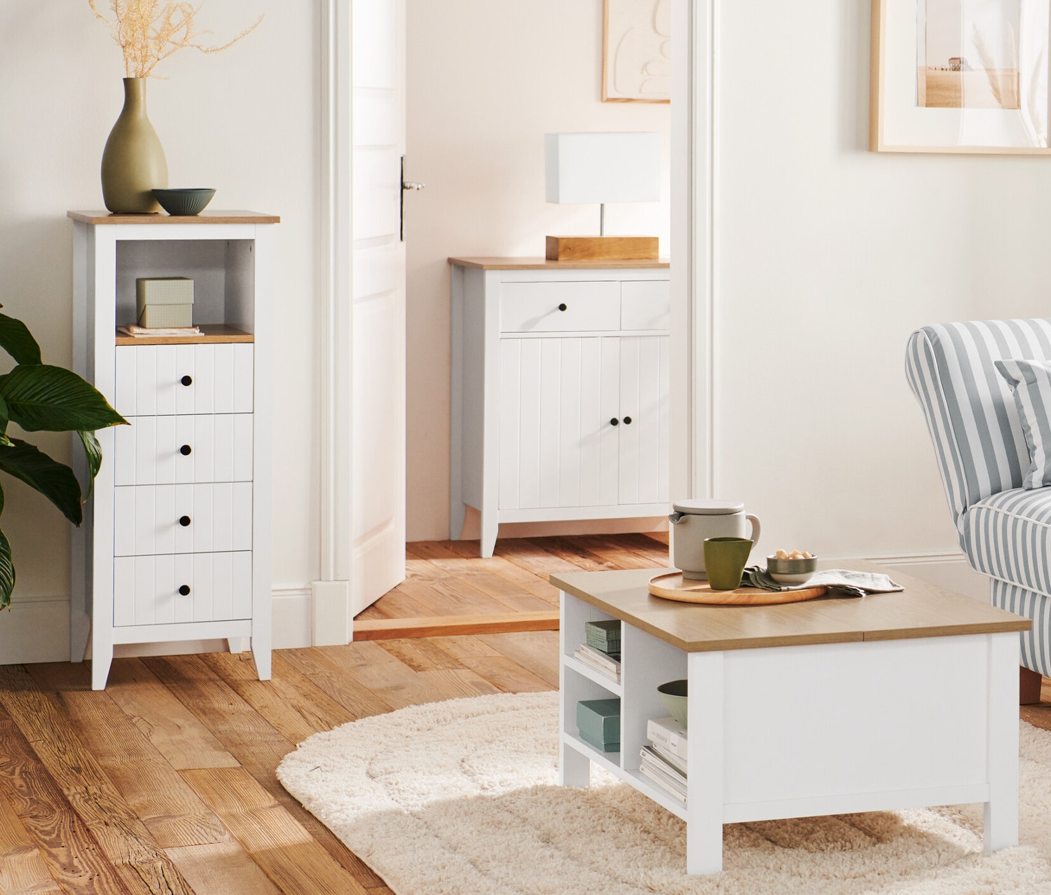 W salonie stoi drewniana lampa stojąca „trójnóg” obok sofy i otwieranego stolika kawowego. W tle komoda z szufladami i lampa stołowa z drewnianą podstawą.