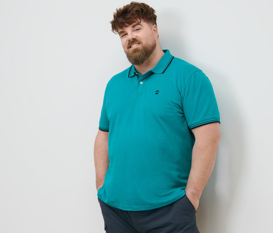 Mężczyzna o brązowych włosach stoi z rękami w kieszeniach granatowych szortów bojówek. Ma na sobie morską koszulkę polo o splocie pique.