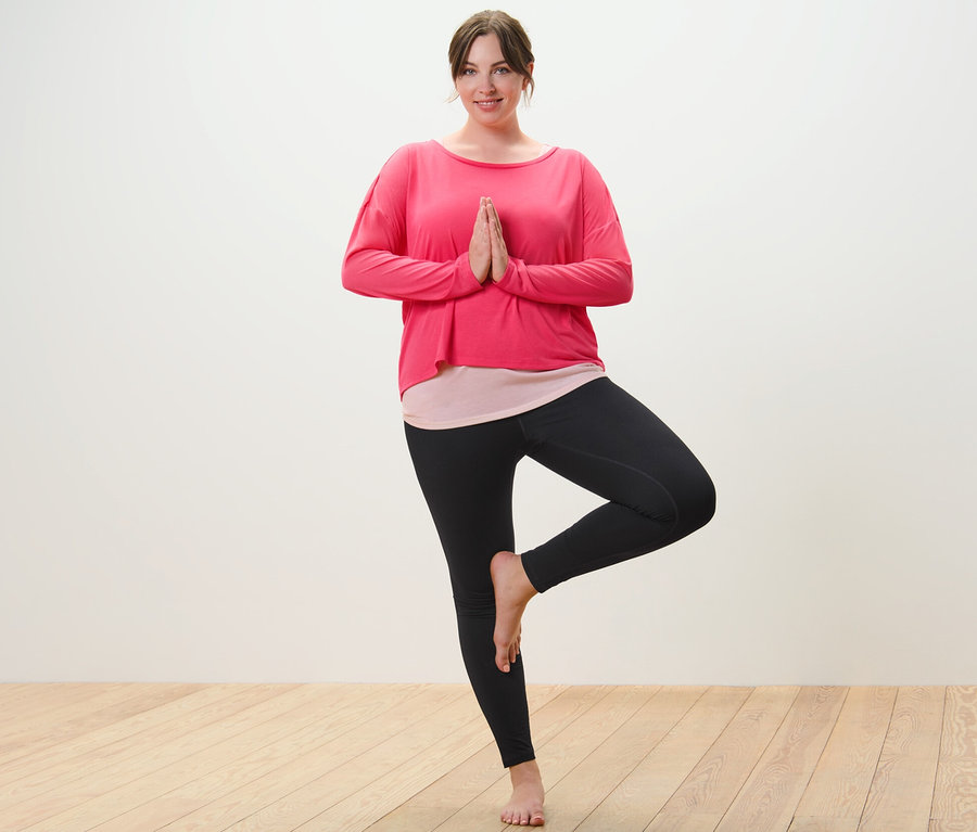 Kobieta w różowej koszulce sportowej 2 w 1 i czarnych legginsach sportowych stoi w pozycji drzewa.