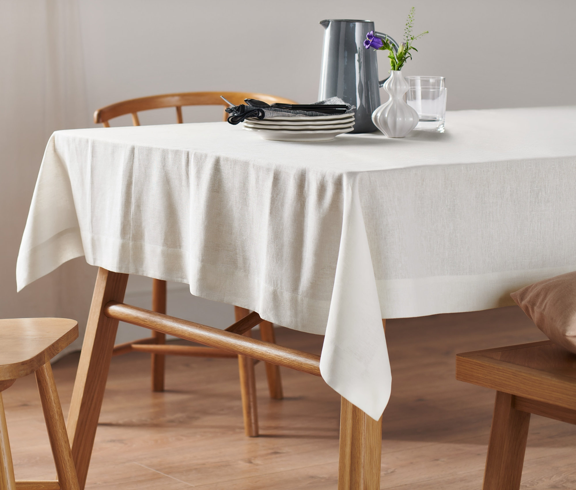 Biały, lniany obrus o standardowej wielkości leży na drewnianym stole. Na obrusie stoi wazon i naczynia.