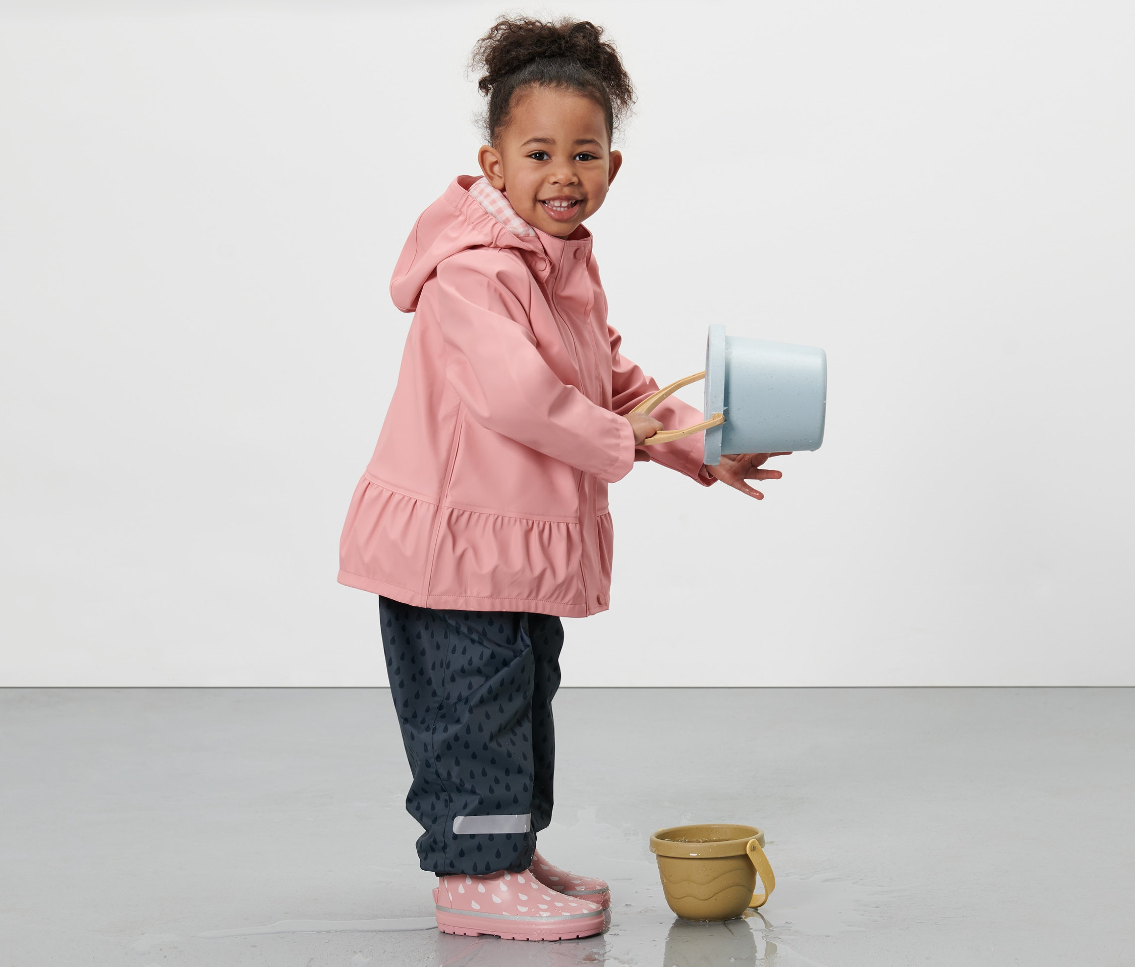 Dziewczynka w kurtce przeciwdeszczowej trzyma niebieskie wiaderko i stoi obok żółtego wiaderka.