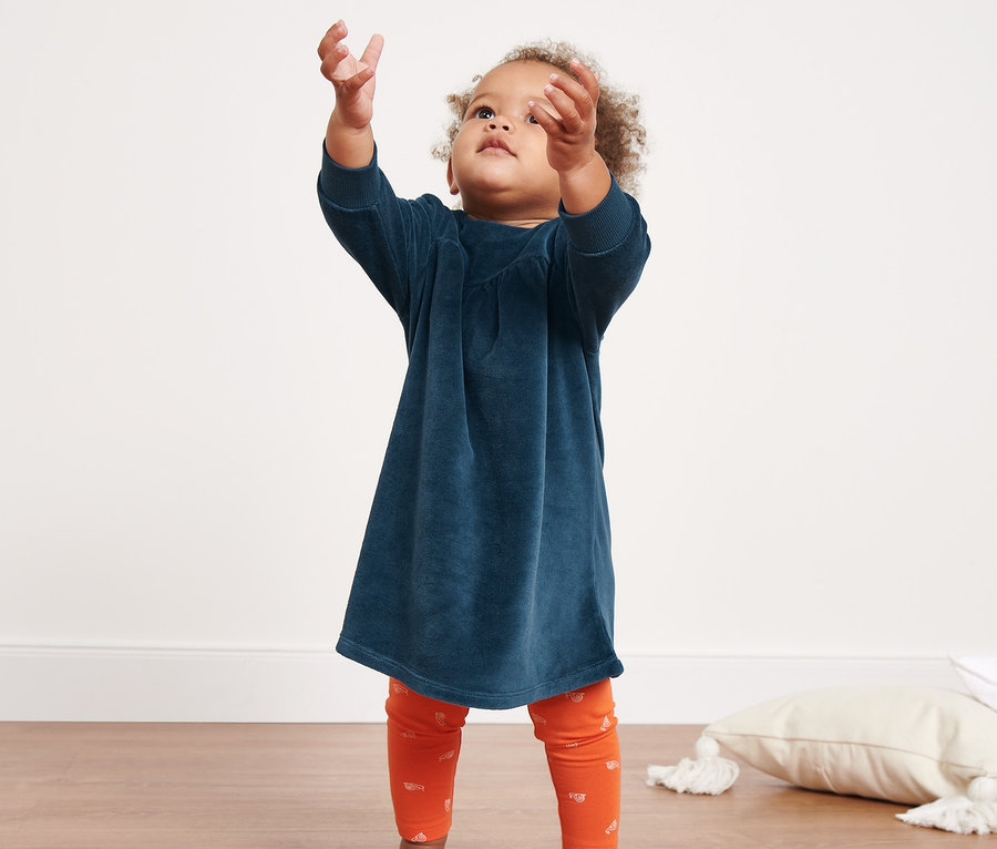 Dziewczynka sięga po bańki mydlane. Ma na sobie sukienkę welurową i pomarańczowe legginsy.