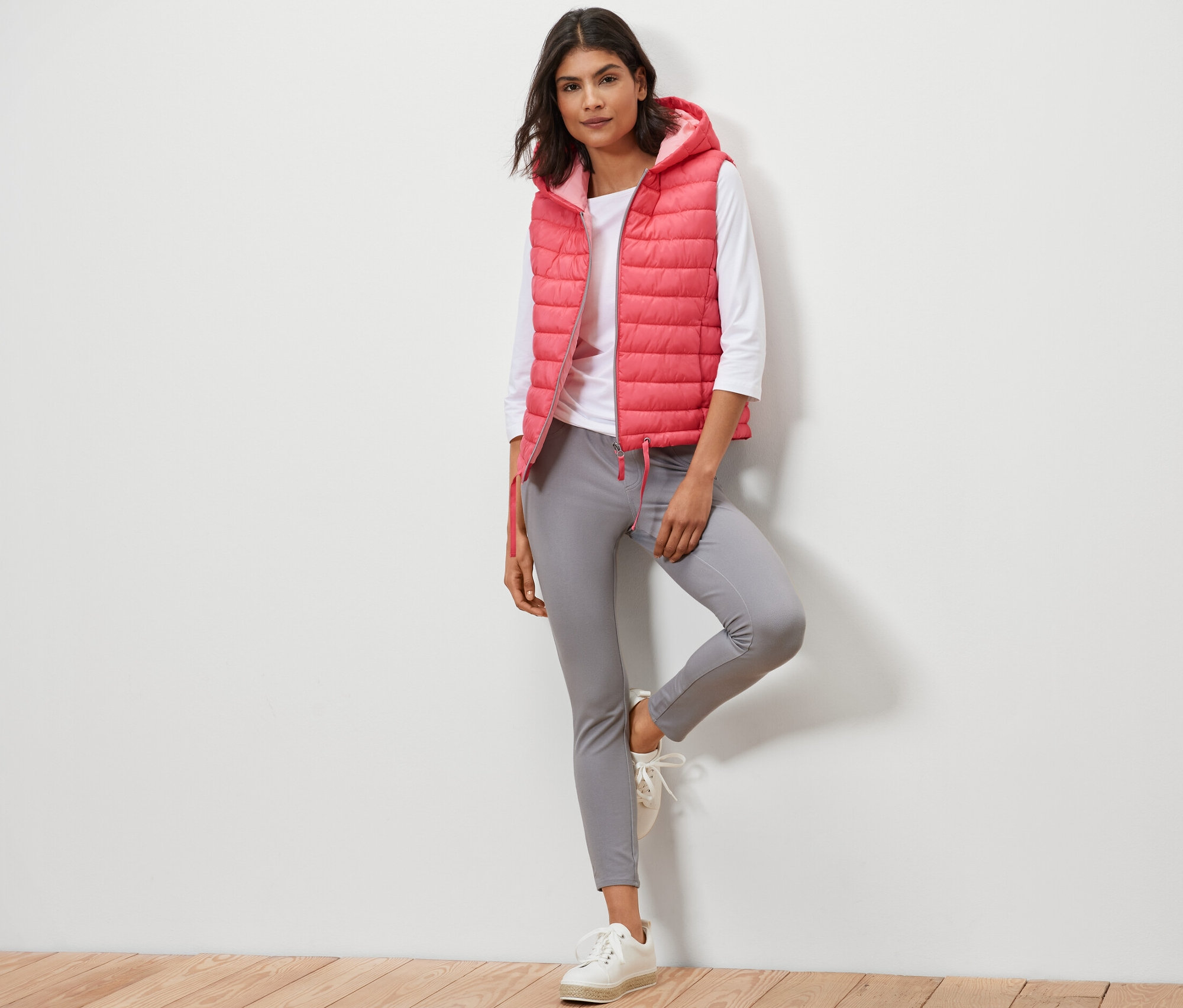 Kobieta pozuje w kamizelce pikowanej z kapturem, białej koszuli i szarych spodniach.