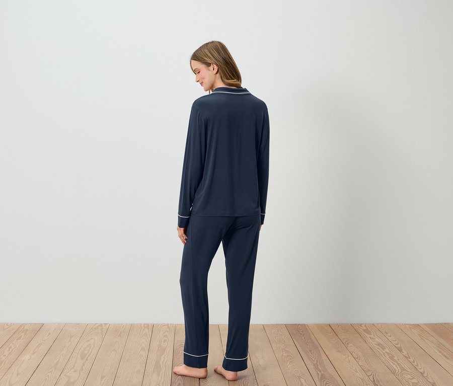 Kobieta stoi tyłem, ubrana w granatową piżamę z białymi lamówkami.