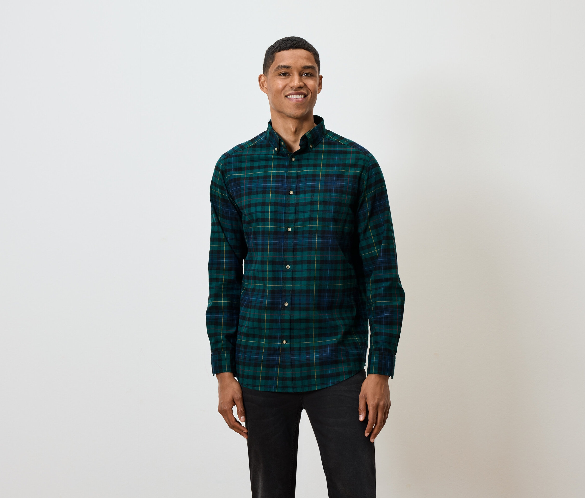 Mężczyzna z krótkimi włosami pozuje na białym tle, ubrany w zielono-niebieską koszulę w kratę z długim rękawem i czarne dżinsy.