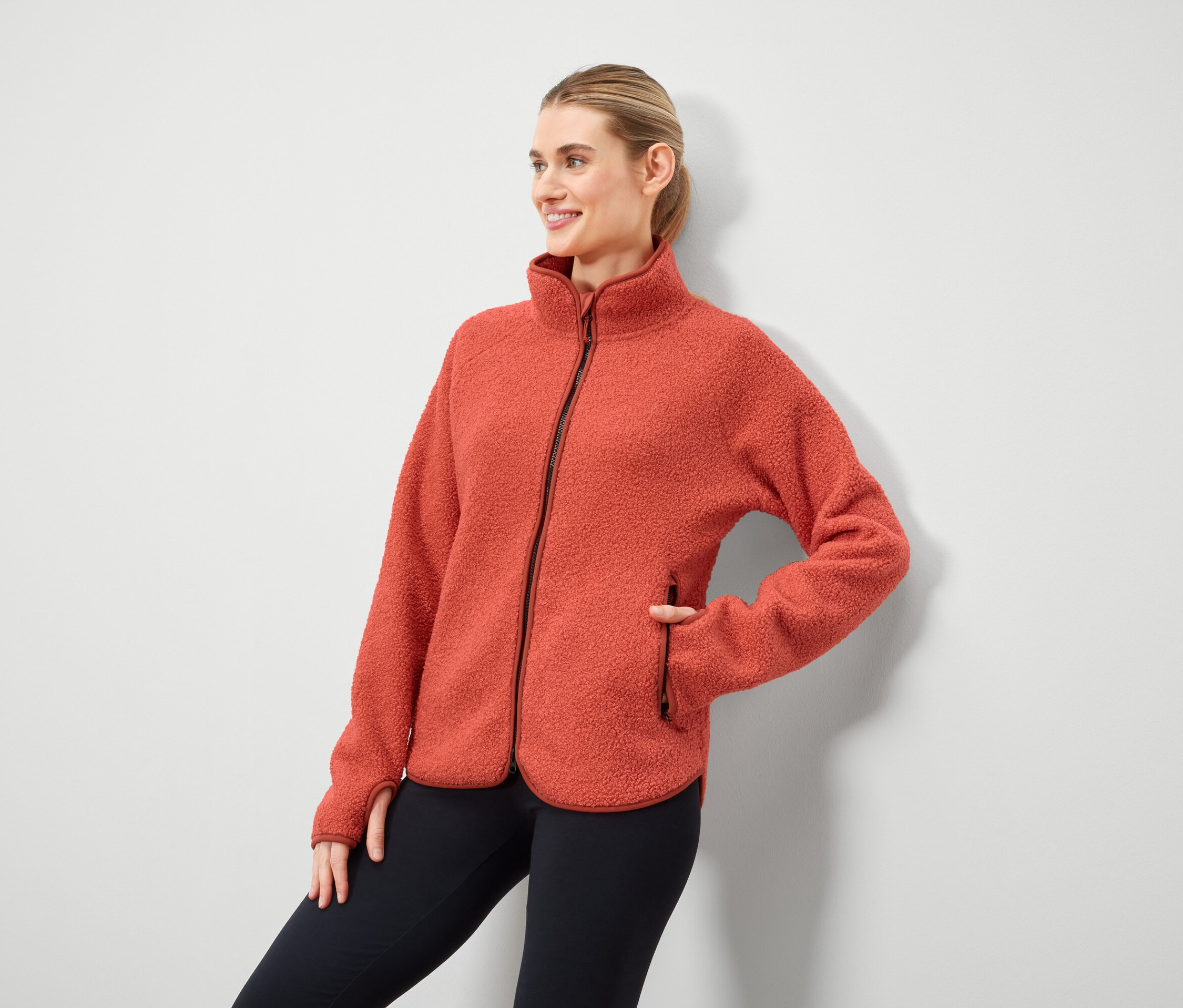 Kobieta pozuje w pomarańczowej polarowej kurtce zapinanej na zamek i czarnych legginsach.