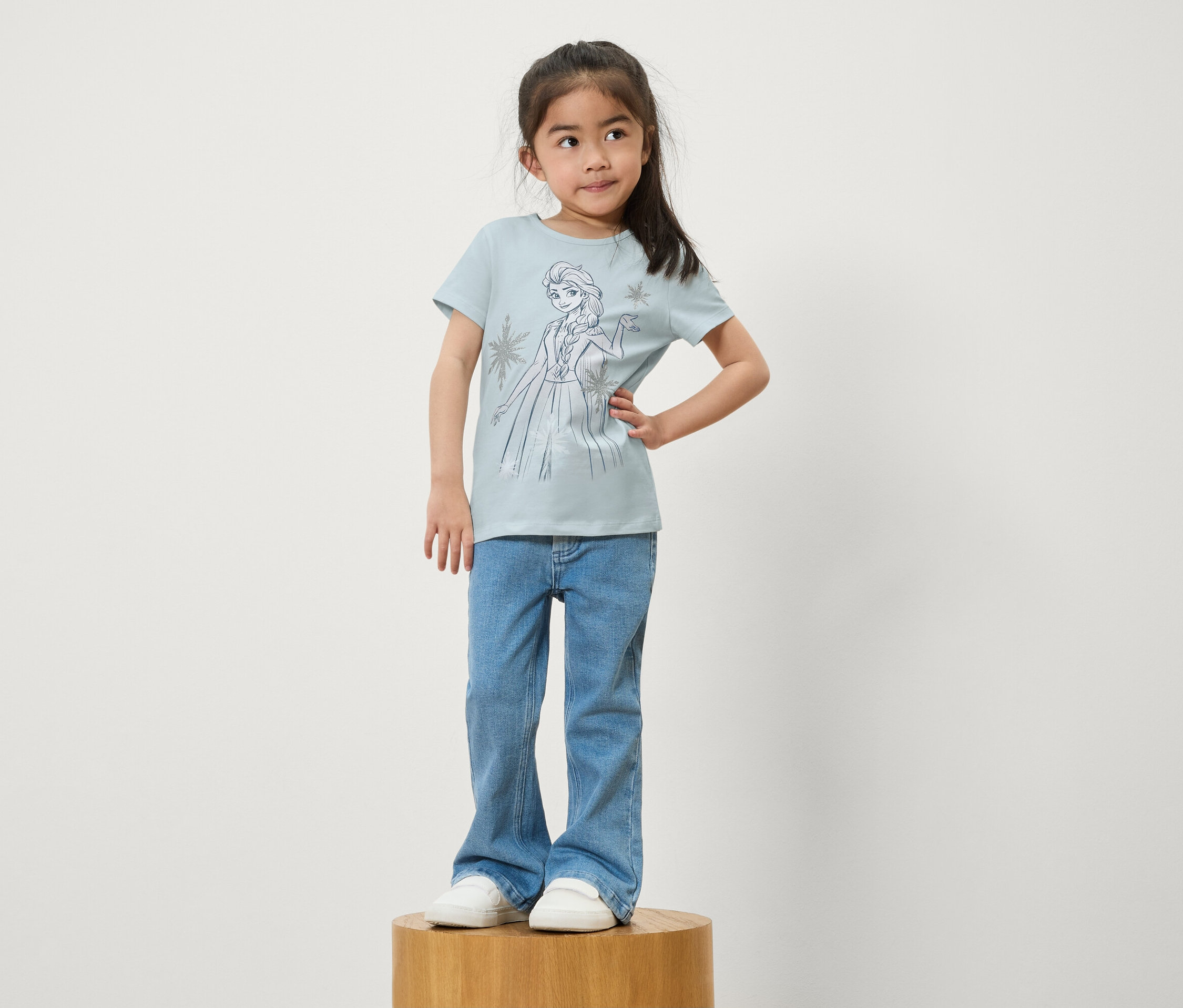 Dziewczynka stoi na drewnianym cokole. Ma na sobie niebieskie dżinsy i jasnoniebieską koszulkę z nadrukiem Elsy z Krainy Lodu.