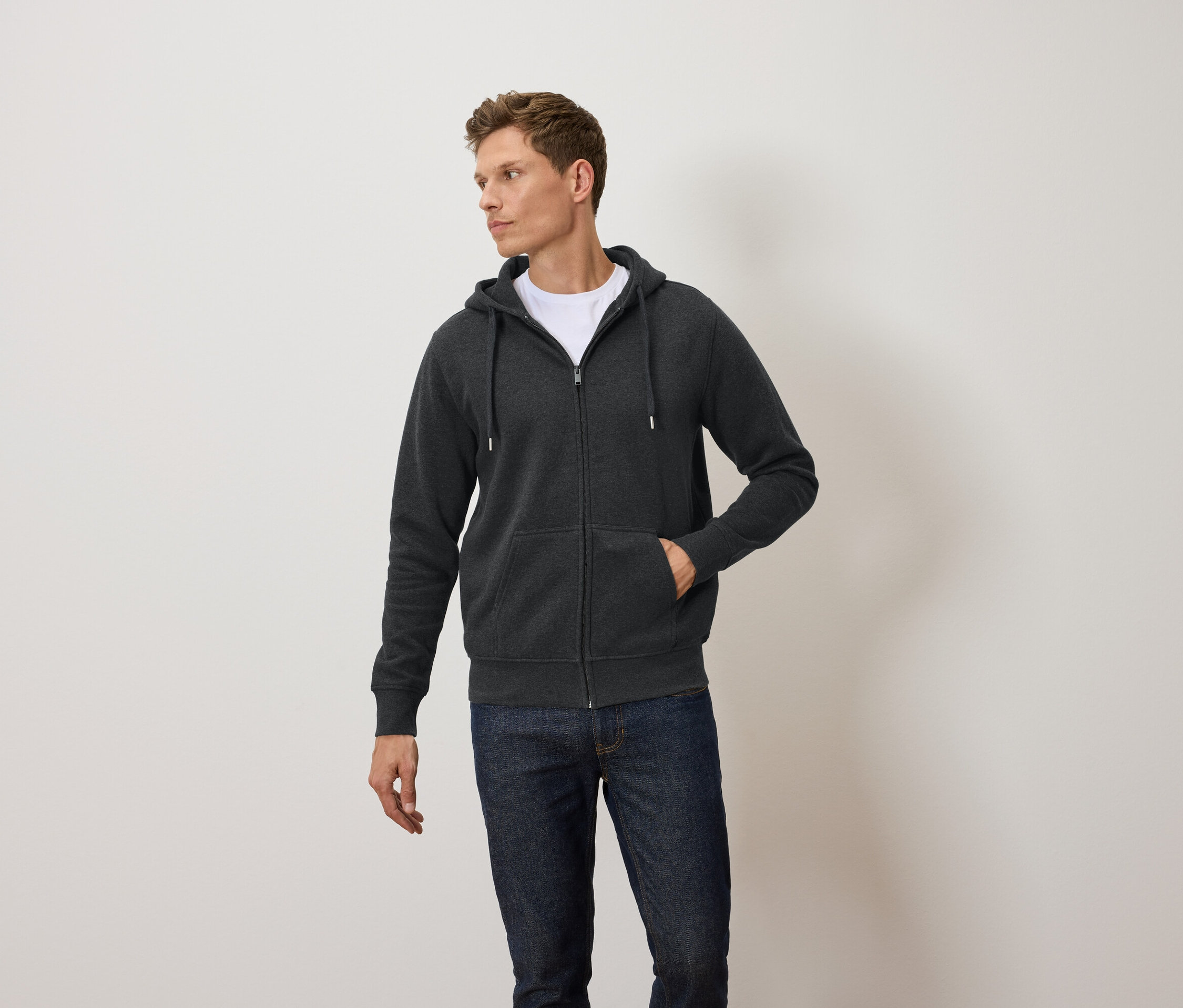 Mężczyzna ma na sobie ciemnoszarą bluzę z kapturem zapinaną na zamek, białą koszulkę i niebieskie dżinsy.