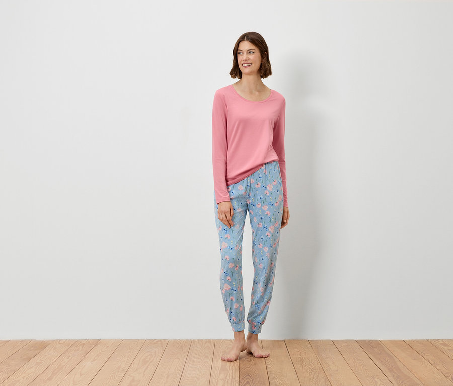 Kobieta stoi, ubrana w różową bluzkę z długim rękawem i niebieskie spodnie w kwiatowy wzór.