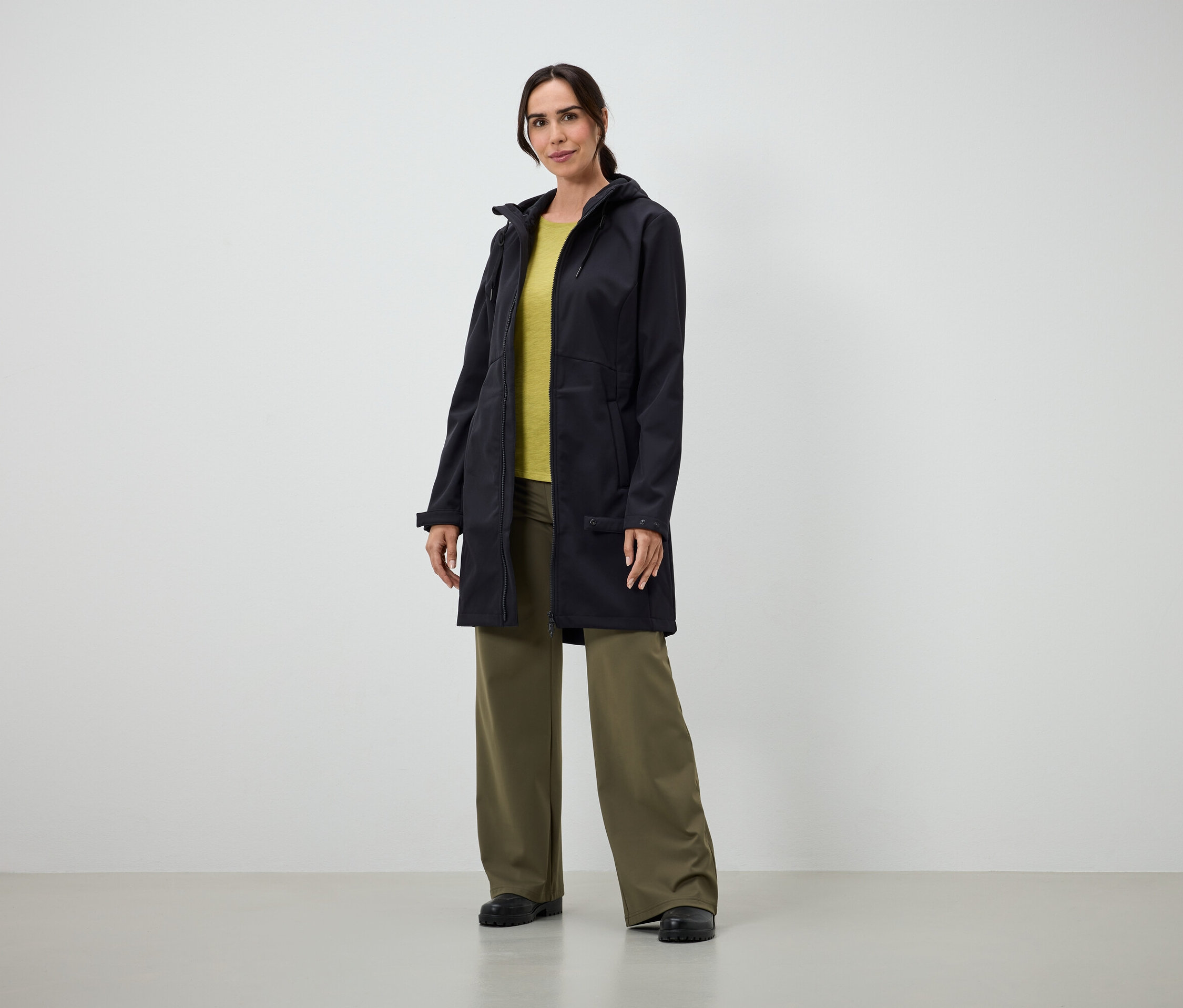 Kobieta w długim czarnym płaszczu, żółtej koszulce i szerokich zielonych spodniach stoi w pomieszczeniu.