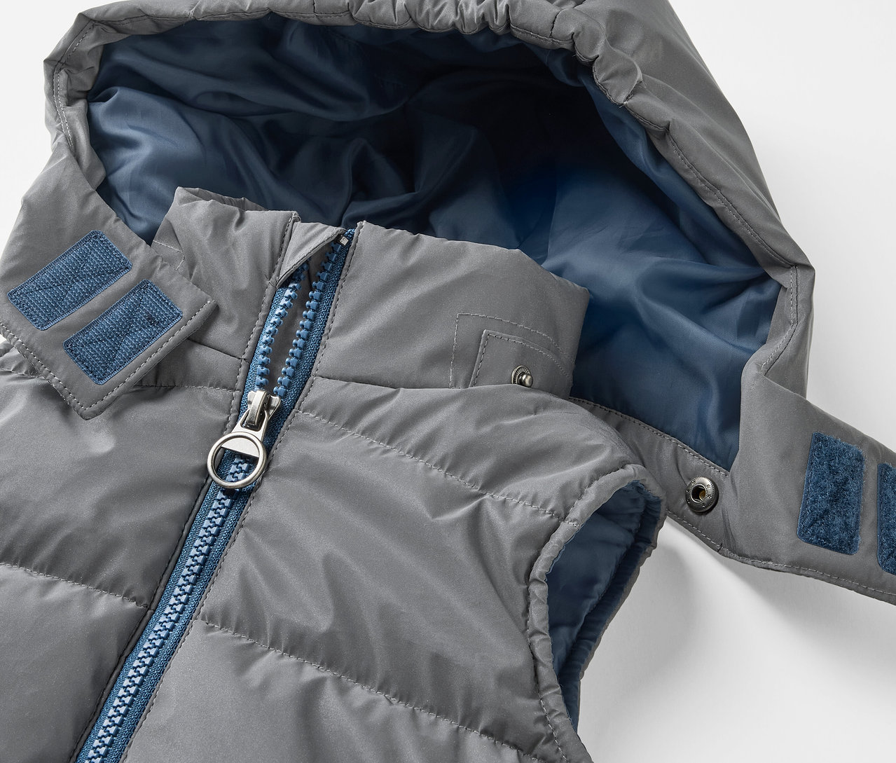 Zbliżenie szarej, pikowanej kamizelki dziecięcej z kapturem. Dziecięca odblaskowa kamizelka pikowana ma niebieski zamek błyskawiczny i niebieską wewnętrzną stronę kaptura.