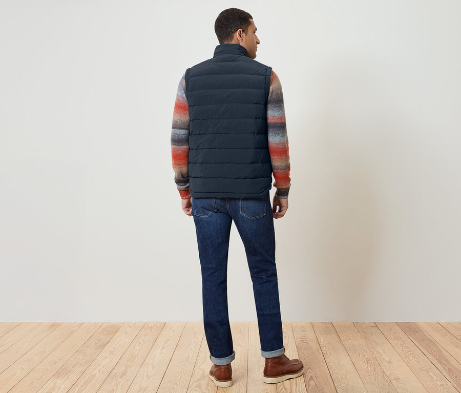 Mężczyzna tyłem do kamery ma na sobie granatową kamizelkę, kolorowy sweter, dżinsy i brązowe buty.