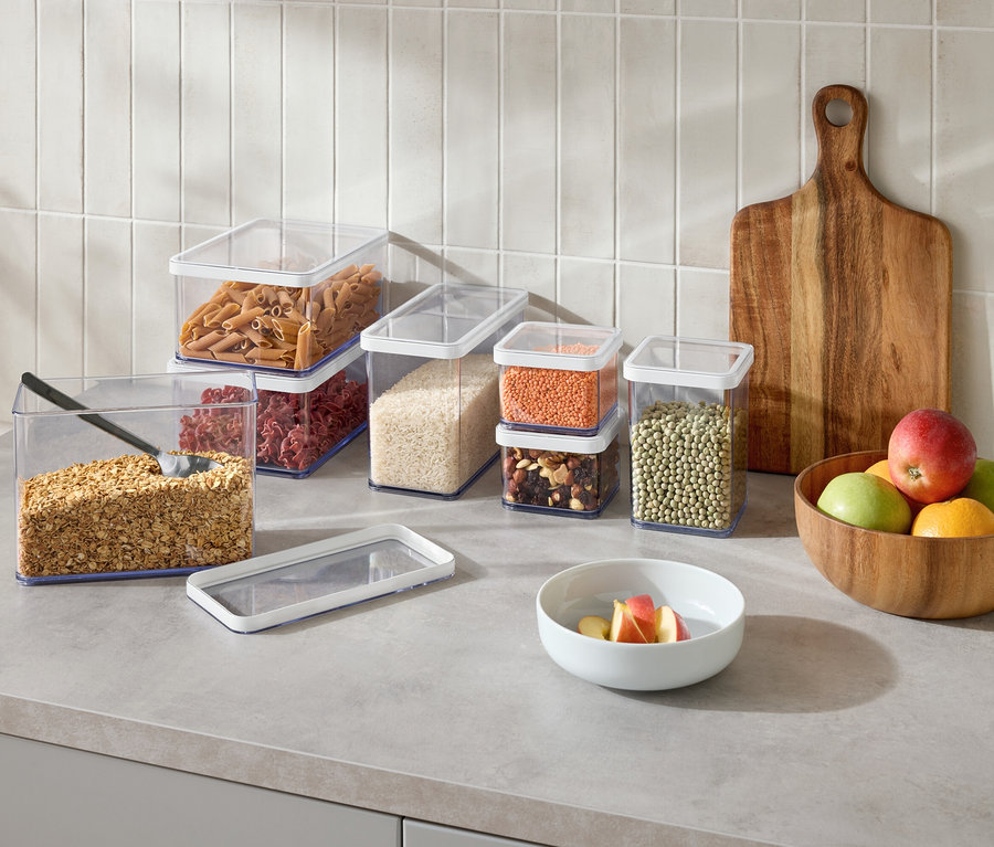 Na blacie kuchennym stoi kilka przezroczystych pojemników do przechowywania żywności z różnymi produktami, drewniana deska do krojenia i miska z owocami.