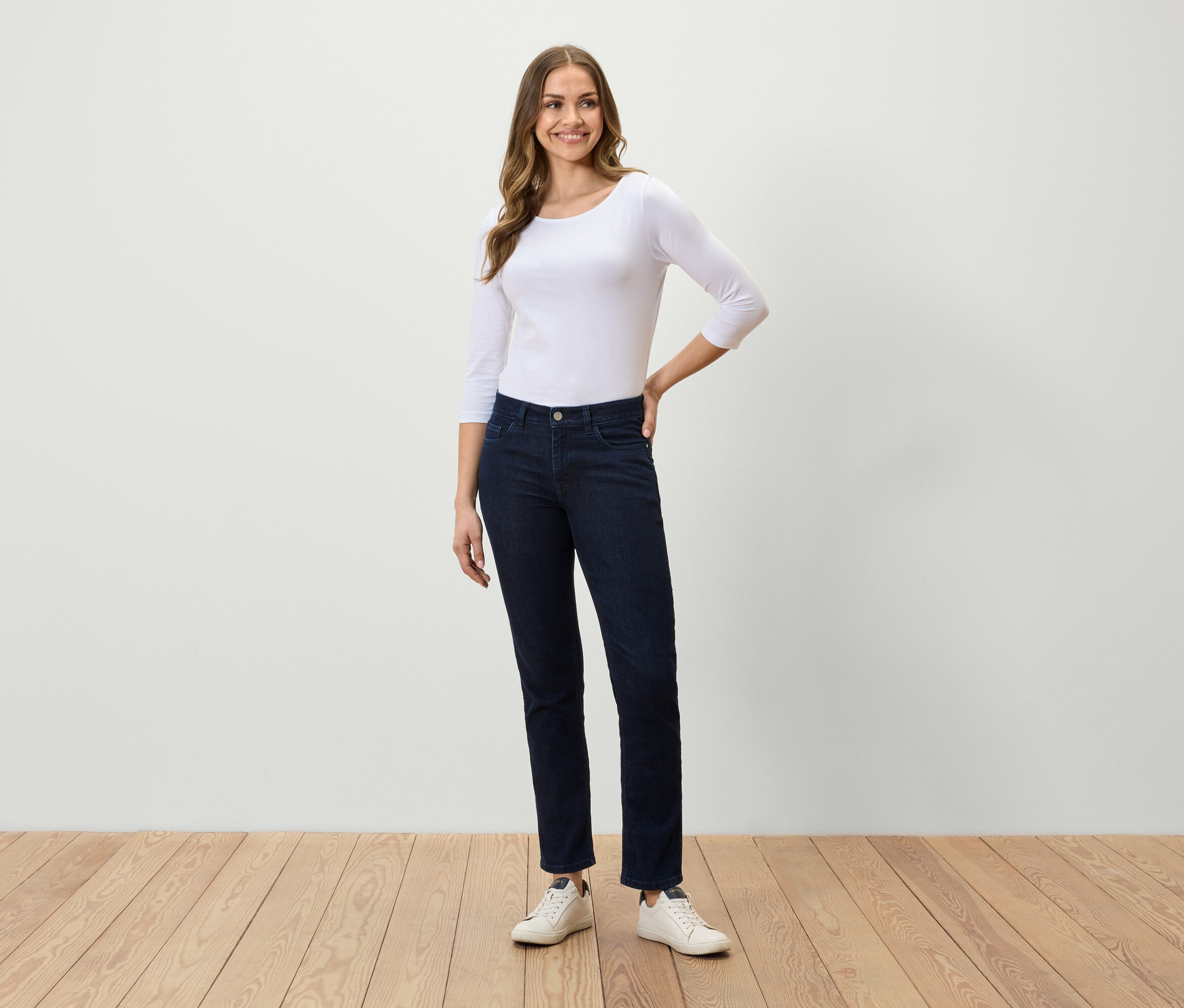 Kobieta stojąca na drewnianej podłodze ma na sobie białą koszulkę z rękawem trzy czwarte, ciemnoniebieskie dżinsy i białe trampki.