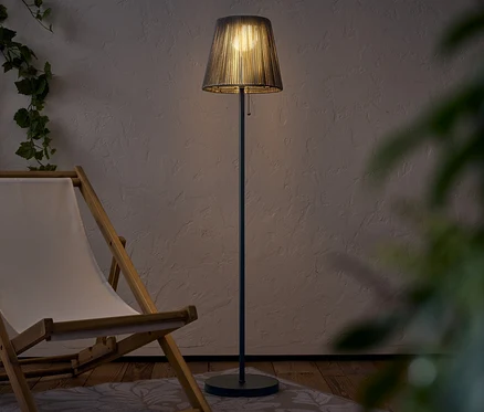 Oświetlona lampa podłogowa z abażurem stoi obok drewnianego leżaka.
