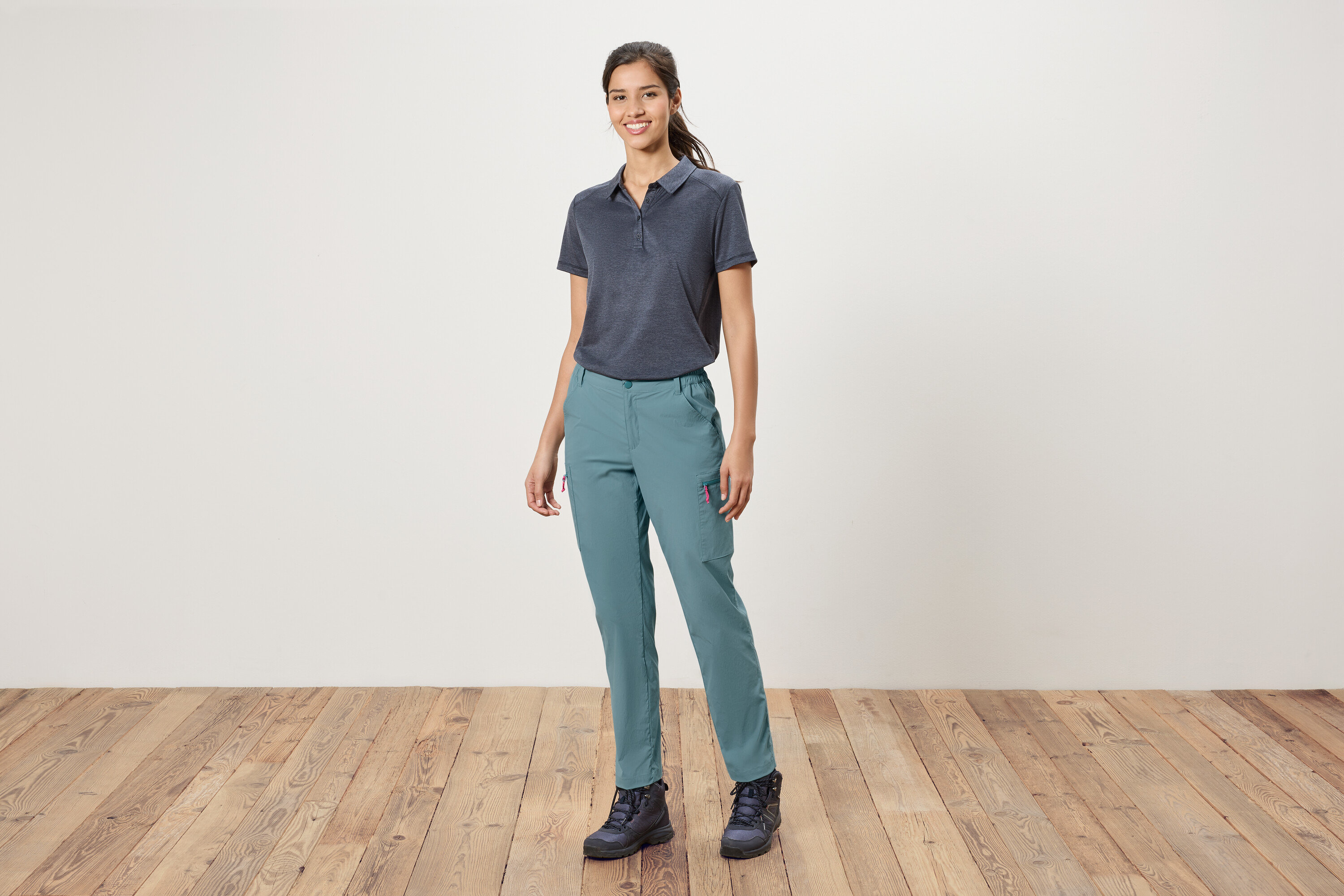 Kobieta stoi na tle białej ściany, ubrana w niebieską koszulkę polo, niebieskie spodnie i buty trekkingowe.