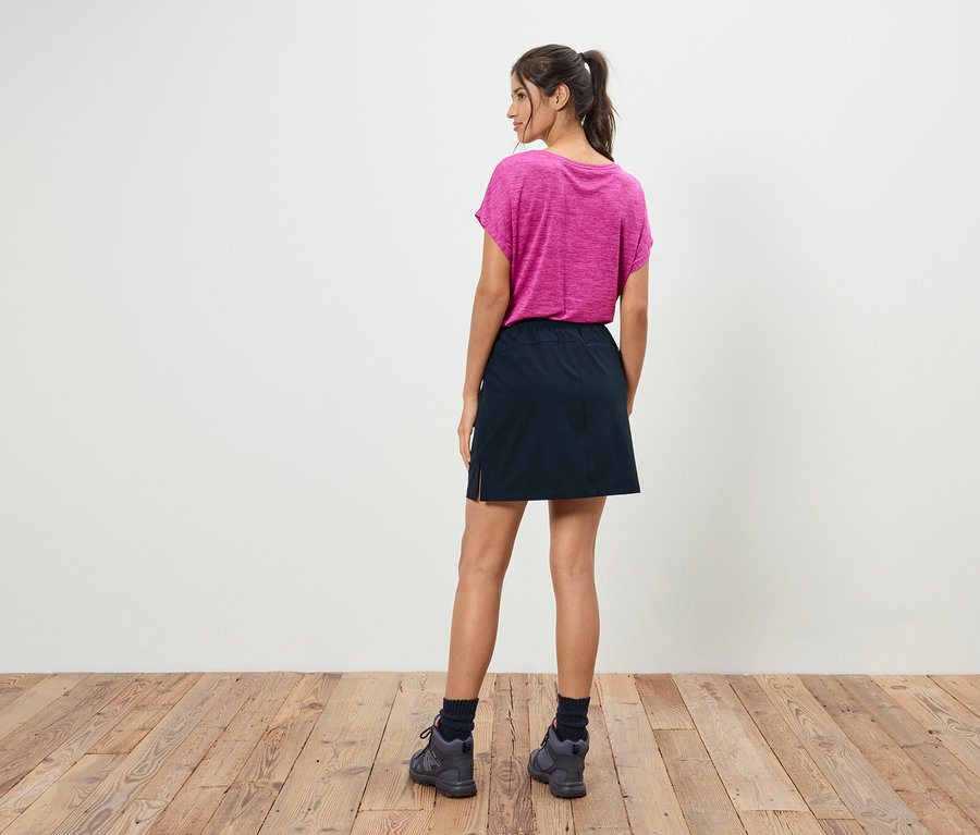 Kobieta z kucykiem, odwrócona tyłem, ma na sobie różowy T-shirt, czarną spódnicę i szare buty.