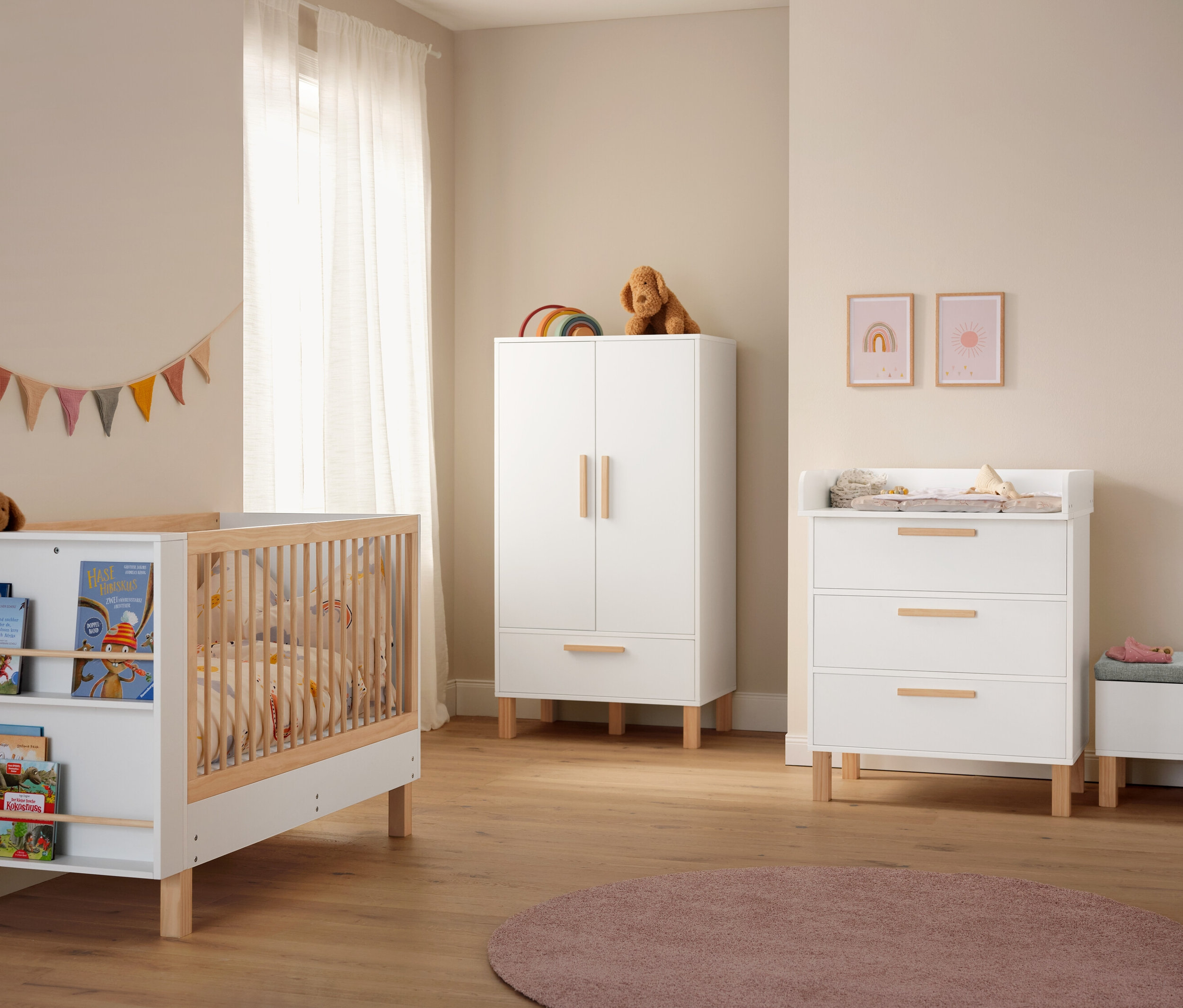 Pokój dziecięcy z białą szafą, przewijakiem, łóżkiem z książkami i różowym dywanem »Charme« o średnicy ok. 160 cm.