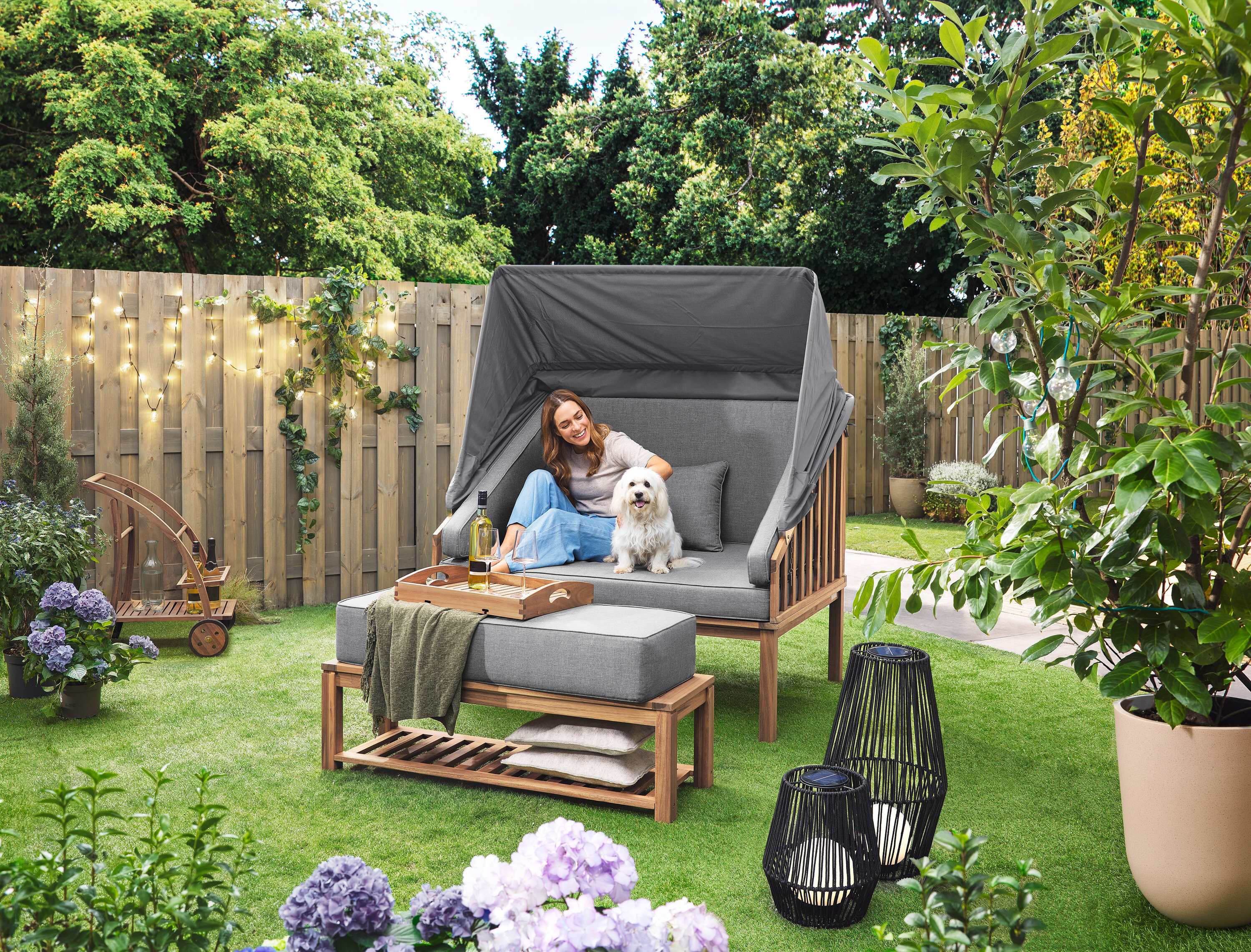 Kobieta siedzi z psem w koszu plażowym Premium Lounge-Strandkorb mit Sonnenschutz-Dach. Na trawniku stoi latarnia solarna ze sznurkiem tekstylnym, duża i latarnia solarna ze sznurkiem tekstylnym, mała. W tle widoczny jest wózek barowy z odłączaną tacą. Na płocie wisi łańcuch LED-Outdoor-Lichterkette mit Netzadapter.