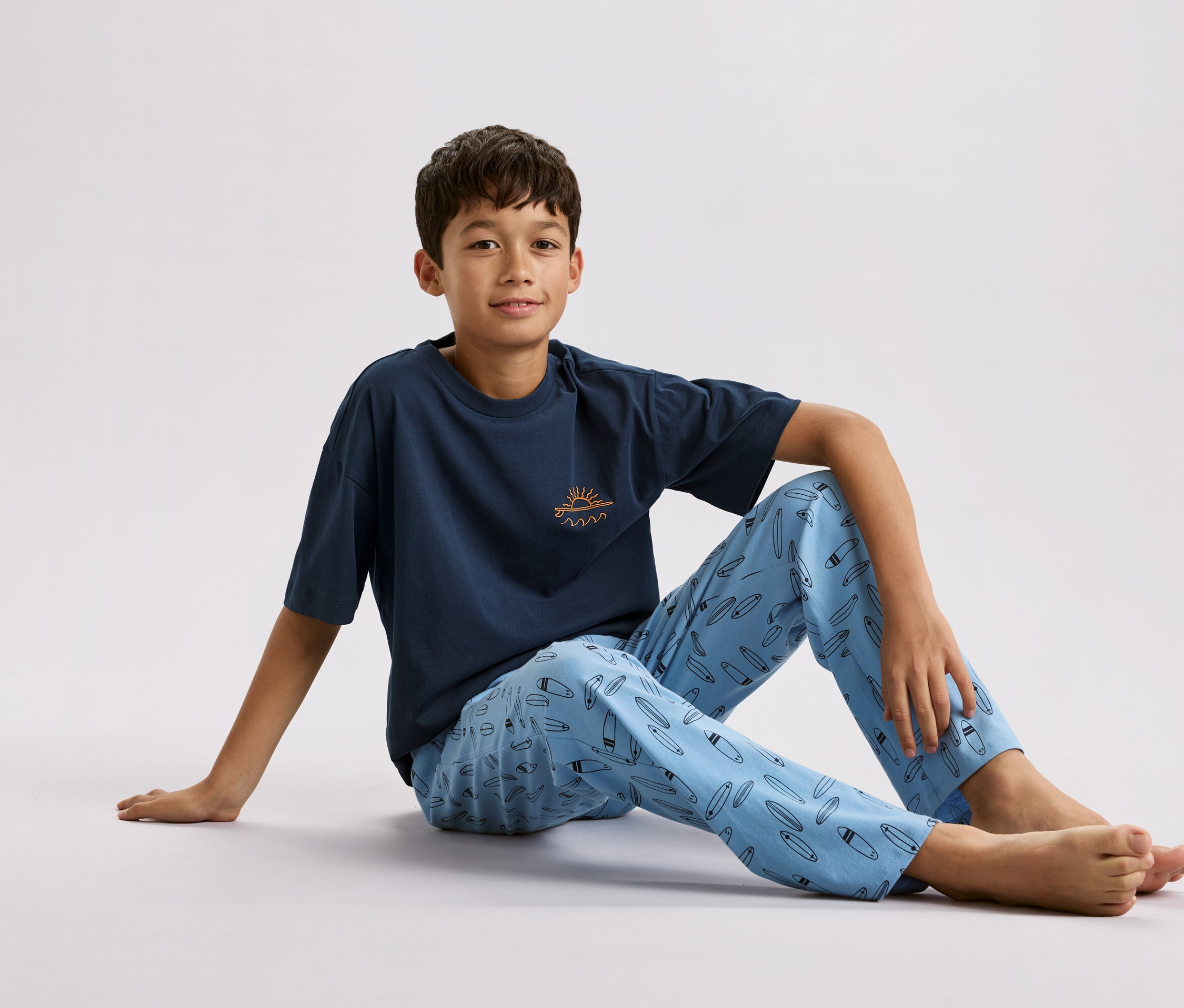 Chłopiec siedzi na podłodze, ubrany w granatową koszulkę z małym słońcem i falami oraz jasnoniebieskie spodnie od piżamy we wzór desek surfingowych.