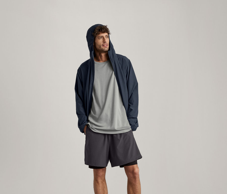 Mężczyzna ma na sobie jasno szarą koszulkę sportową, granatową kurtkę z kapturem i szorty.