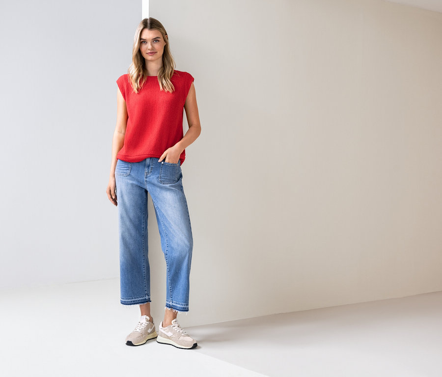 Kobieta stoi w jasnym pokoju, ubrana w czerwoną bluzkę, niebieskie dżinsy i trampki.