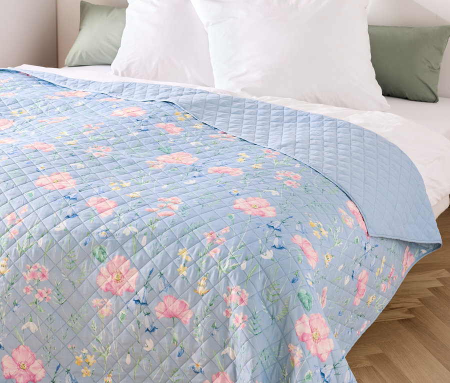 Niebieska narzuta w kwiaty na łóżku z białymi poduszkami.