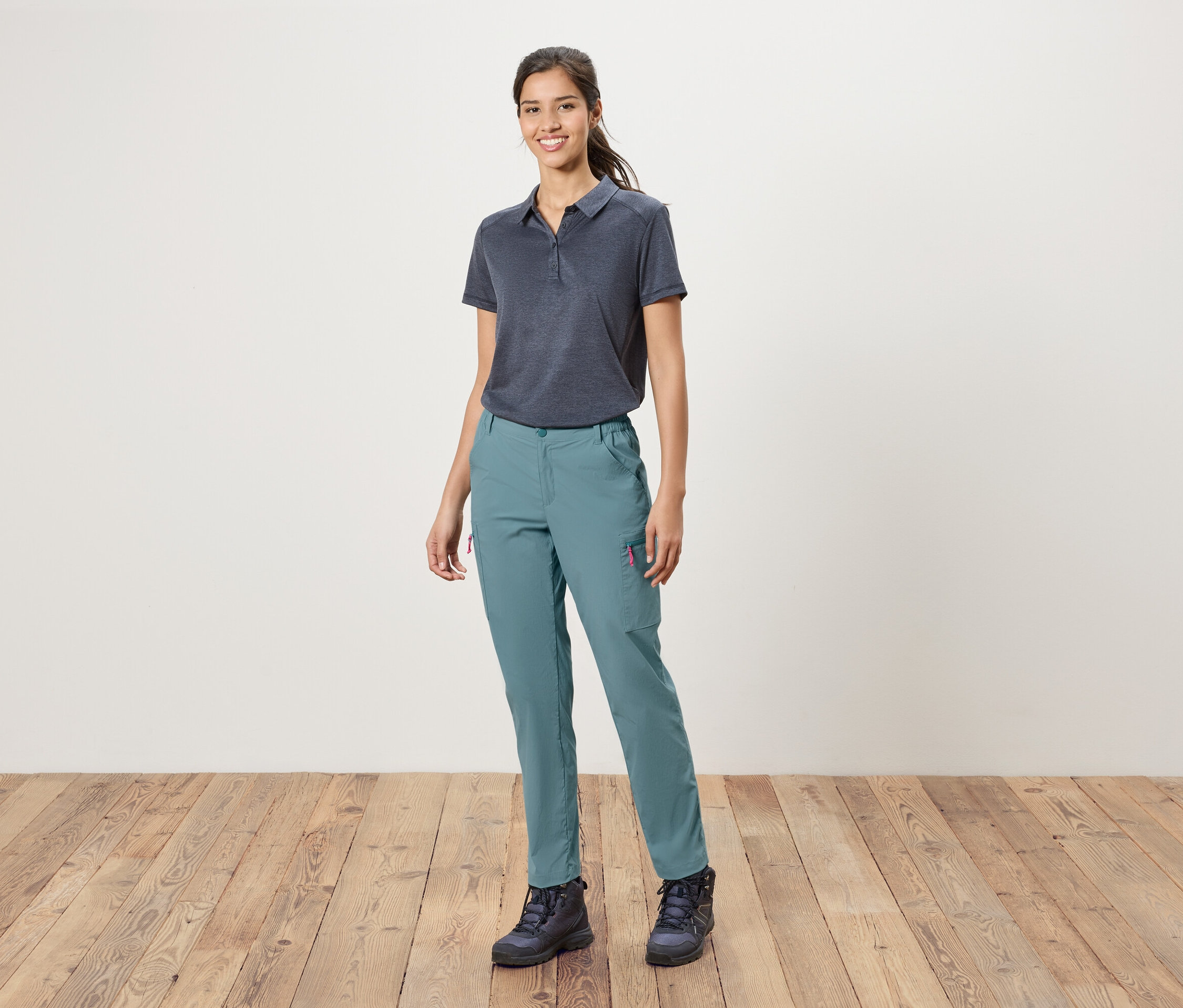 Kobieta stoi na tle białej ściany, ubrana w niebieską koszulkę polo, niebieskie spodnie i buty trekkingowe.
