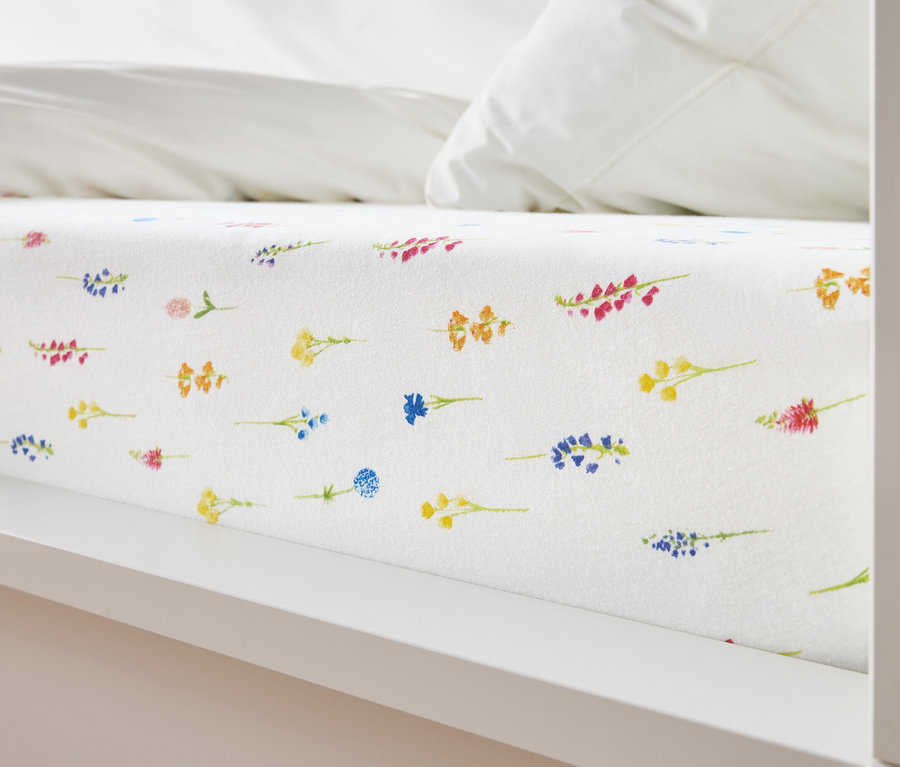 Bliski widok łóżka z białym prześcieradłem pokrytym kwiatowym wzorem w kolorach żółtym, niebieskim, pomarańczowym i różowym.