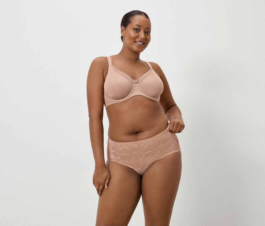 Kobieta o ciemnej karnacji ma na sobie beżowy biustonosz i pasujące koronkowe majtki na tle prostego tła.